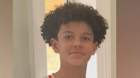 Noah smiling at the camera. He is wearing a white football shirt with red edging and has short curly hair.