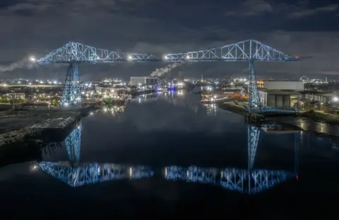 Simon McCabe A metal blue bridge at night reflected in the dark river below it. City lights can be seen stretching into the distance, with smoke from industrial chimneys.