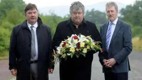 Pacemaker Three men standing side by side as one holds a wreath. From left to right is a man with short brown hair black coat, grey trousers, white shirt and grey tie. then a man with short grey hair and a black coat. Then a man with short grey hair, grey moustache wearing grey jacket, white shirt, blue striped tie and black trousers. Behind them is a road where the attack occured.