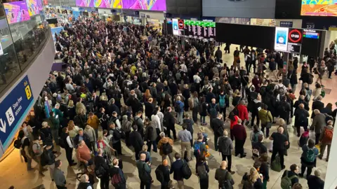 The concourse at London Waterloo pictured from above. It is full of passengers standing in queues and in groups, stretching away to the edge of the frame. Many are looking up at the electronic departures boards. 