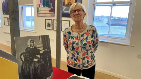 Nina Brown is a woman in her 50s or 60s with short grey hair and glasses and wearing a cardigan covered in blue and orange leaves. She is standing next to a black and white photograph of Mary Ann Macham seated on a chair and through the window behind her can be seen the town of North Shields 