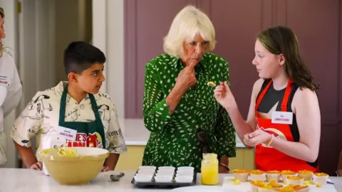 PA Media Queen Camilla stands between two children, aged about 12 or 13, as they bake cupcakes. The Queen is wearing a green spotted dress and has the tip of her finger in her mouth, licking curd off of it. The two children are looking at her. There are cooked cupcakes, a mixing bowl, a jar of lemon curd and filled cupcake cases ready to be put in the oven in front of them.
