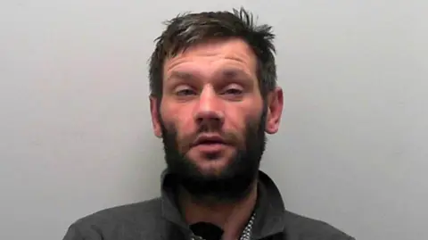 A custody picture of a man with short hair, facial hair, who wears a dark collared shirt.