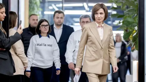 EPA Vêtue d'un costume beige et de cheveux bruns courts jusqu'au menton, la présidente de la Moldavie marche devant son entourage, tenant une feuille de papier au format A4 dans sa main droite.