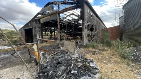BBC/Suzy Lawson A barn which has been destroyed by a fire. There are holes in the roof and the sides of the building. There are also charred remains in front of the barn.