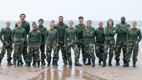 Pete Dadds | Canal 4 Os celebridades recrutam, usando fadigas militares, alinham -se em uma praia com as costas para o mar. Eles estão em duas fileiras, alguns com os braços cruzados, com expressões sérias.