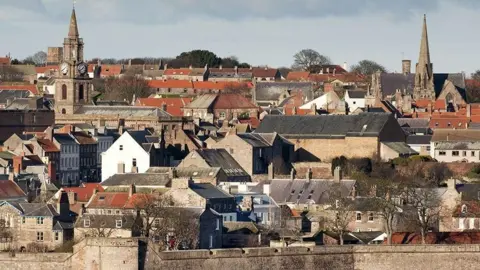 English Heritage Berwick, viewed from outside the town walls. Old stone houses, are jumbled together, some with red tile roofs and some with black slate. It is a sunny day with the sun bouncing off the occasional white gable end, but the trees have no leaves. There are two church spires, one on either side.