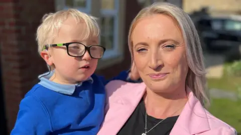 BBC Head and shoulders shot of five-year-old Harvey Hind with blonde hair wearing black glasses and royal blue school sweatshirt over a pale blue polo shirt. He is being held by his mum in the garden of their house in the sunshine. She has long straight blonde hair and is smiling and wearing a pale pink blazer over a black tshirt