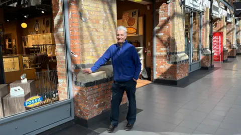 Andy Binks stands in a shopping centre next to his old work bench. He has white hair and is leaning next to the spot. He's wearing a royal blue fleece and dark, navy work trousers. 