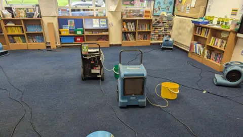 A picture of a classroom showing several dehumidifiers can be seen helping to dry out a sodden blue carpet. Bookshelves and wall displays can be seen in the background.