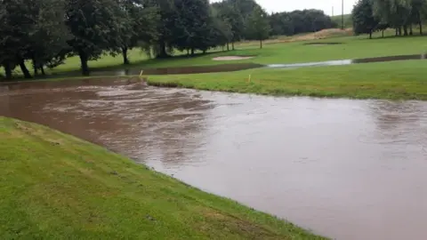 A flooded golf course. There are trees in the distance and bunkers. There is a large body of water in the centre of the image which is brown in colour.