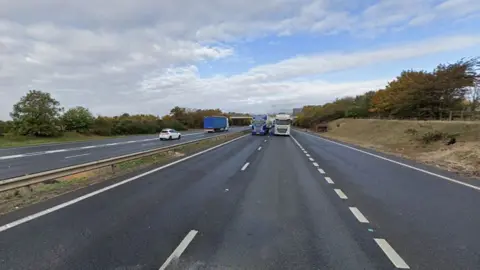 The M11 viewed from the right carriageway, with the central reservation. This is a google image from a different date so no lanes are closed. Two lorries side by side are coming towards the camera, while a white car and lorry are on the other side. 