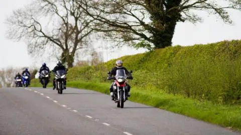 Getty Images Bikers are riding on a main road that goes through countryside in the generic image. They are passing a hedgerow with a grassy verge and there are trees in the fields behind them.