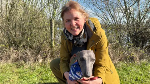 A greyhound wearing a blue bandana with the branding 'Ambassadog' is cradled by his owner who is a woman wearing a mustard coloured coat and a floral scarf. They are crouched on grass with bushes and a lake in the background.