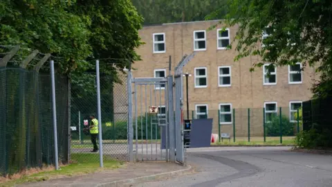PA Media The photograph shows the entrance to a secured former military site.
A metal security gate stands open across a tarmac access road. The gate is surrounded by chain-link fencing topped with angled extensions carrying strands of barbed wire. The road curves gently to the right beyond the entrance.