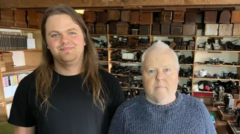 Gabriel has long hair and it standing next to his father with grey hair and hundreds of sewing machine paraphernalia in the background.