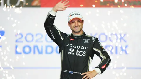 Bradley Collyer/PA Wire. Mitch Evans of Jaguar TCS Racing celebrates finishing third in the championship following the Hankook London E-Prix, ABB FIA Formula E World Championship, Round 17 race at the ExCeL London. He is smiling and waves with his right arm.