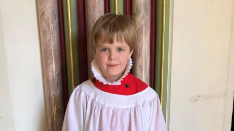 Supplied George smiles at the camera wearing white and red chorister robes. He has blond hair, some of which hangs over his forehead.