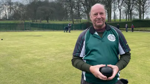 A man with short grey hair stands on a bowling green holding a bowling ball. He is wearing a green and black polo shirt over a green long-sleeve T shirt.