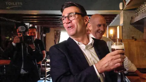 PA Media Andy Burnham holding a pint of beer at a bar, while looking across his shoulder to a person out of shot while talking. There is a camera crew and his new chancellor John Healey behind him. Burnham has short black hair and thick framed glasses and he is wearing a suit jacket over a white shirt. Healey, who has a bald head and is wearing a white shirt, is smiling.