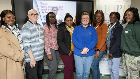 The Royal Wolverhampton NHS Trust Seven women and one man posing for a photograph in a line. All those pictured are smiling at the camera. They are standing in a room with a green carpet. A pull-up banner saying HOPE is behind them to the right. On the left behind them is a digital screen with an image of a smiling young women and words partially obscured, but Your NHS can be made out.