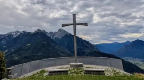 Submitted A large cross on top of a raised platform has a grey barrier in front of it. It overlooks several mountain tops on a cloudy day.
