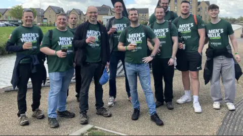 Ten men standing by a small river with a housing estate in the background. They are all smiling to camera, wearing green T-shirts with a logo saying Men's Walk and some are holding pints of beer.