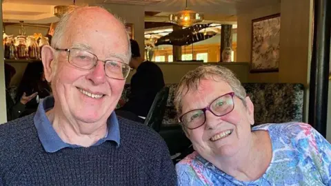 Laurence Neal Laurence Neal and Jill Neal together in a pub or cafe, both smiling broadly. Laurence is on the left wearing a mid blue shirt under a dark blue jumper. He has white hair and is wearing glasses. Jill is on the right and is leaning towards him, wearing a blue, green and pink top. She has short grey hair and is wearing glasses. 