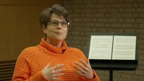 Composer Emily Howard sits wearing an orange jumper in front of a music stand gesticulating with her hands. 