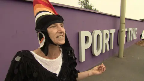 Rachael Kurtz, wearing a puffin costume, which includes a tall, orange "beak-style" headdress. She is gritting her teeth and gesturing passionately with her left hand.