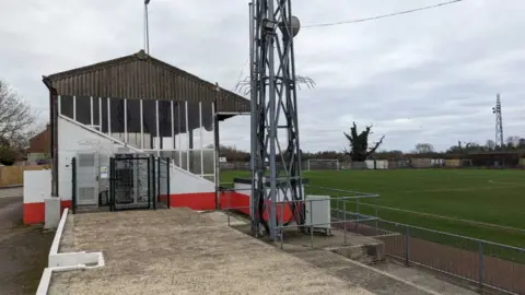 Part of the ground at Stag Meadow, with a covered terrace to the left of the picture and the grass pitch to the right, with the bottom part of a floodlight in the centre of the picture.