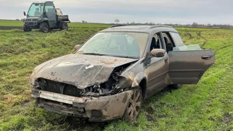 A dark-coloured car caked in mud, its front bumper hanging off, abandoned in a field in the Fens.
