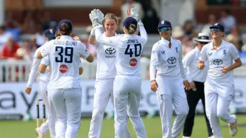PA Media A group of female cricketers, all dressed in white, high five each other on a cricket pitch.