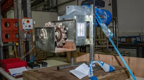 HSE Metal machinery that contains cogs and is is fixed to a bench. Blue and white police tape is tied to the bottom of the machine, which also has a police incident tag also attached to it.