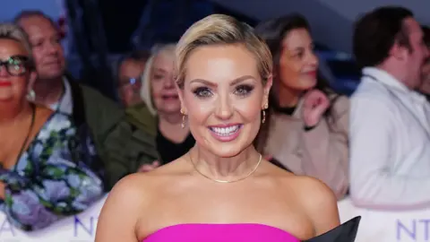 PA Media A woman in a pink dress smiles towards the camera, behind her is a crowd of people