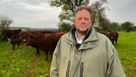 A man with short grey hair and a grey beard stands in a field, wearing a light brown coat. Behind him are several brown cows, including a calf.