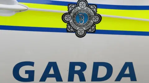 Garda is written in large blue letters underneath a blue and yellow stripe along a car door. The gardaí badge is above this and is made up of Celtic designs with a blue centre.