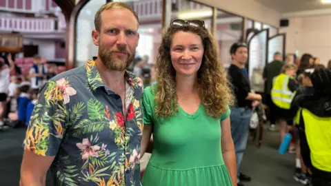 Matt and Sarah Winter are smiling and have their arms around each other. Matt is wearing a colourful black shirt with plants and flowers and Sara is wearing a green dress.