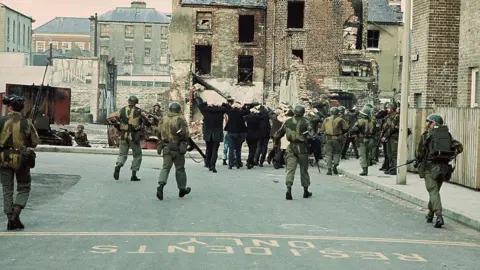 Getty Images Soldiers armed with guns and camouflage clothing and hard hats march arrested civilians with their hands behind their head down the street. The buildings around them are partially knocked down.
