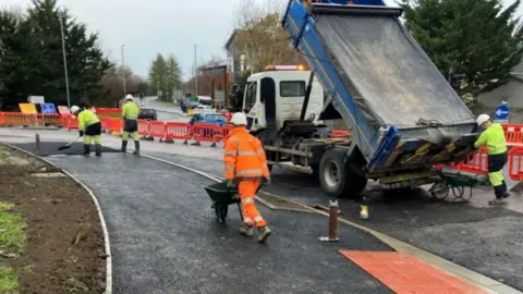 Wiltshire Council People in high-vis jackets with trucks and red fences with signs in the distance.