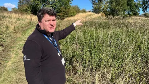 BBC A man with short brown hair and wearing a black quarter zip jumper and a blue lanyard is stood in a field outside, surrounded by trees and bushes. 