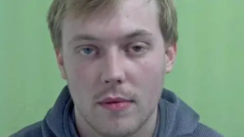 IOM CONSTABULARY Police mugshot of Lewis Kelly, who has short blonde hair and is wearing a grey hooded top. The background behind him is green.
