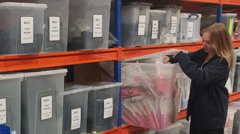 Little Stars Shropshire A volunteer stands next to boxes of boys' black and grey school trousers.