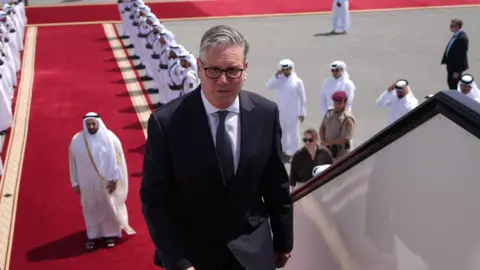 Keir Starmer boarding a plane at the airport in Doha, Qatar. There is a red carpet, lined with men, leading up to the plane