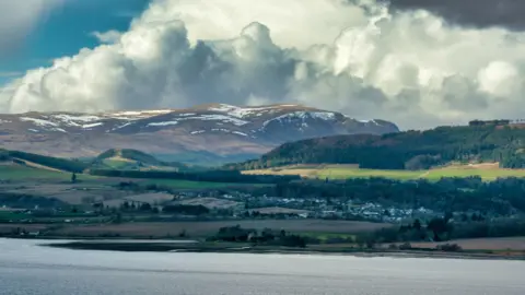 Houses and other buildings of Dingwall below rolling, tree-covered hills and, in the background, the Munro Ben Wyvis. In the foreground is the Cromarty Firth.