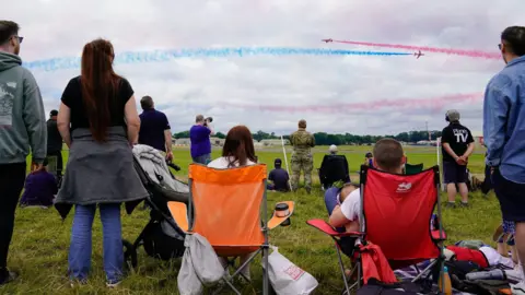 People are sitting on camping chairs and standing on grass watching a flight display. The aircraft are colouring the sky in red and blue.