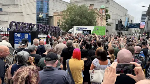 Hundreds of people crowded around the Black Sabbath bench in Birmingham. A purple floral arrangement reading 'Ozzy Forever' is visible above the bench.
