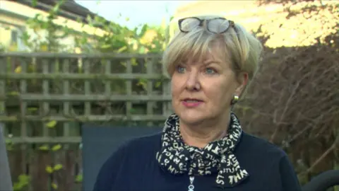 BBC Pauline Shepherd has short blond hair and blue eyes and is wearing glasses on top of her head, a black scarf with white letters on it and a navy top
