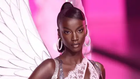 Getty Images for Victoria's Secret Anok Yai poses, wearing a lacy top, hoop earrings and wings attached to her back.