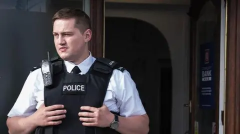 Finnian Garbutt, a man with dark hair, wearing a white shirt with black tie and a black support vest with the words police on it. He is wearing a black watch on his left wrist and there is a brown wooden door in the background.